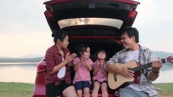 família feliz aproveitando a viagem nas férias de verão. mãe e filho sentam-se no porta-malas do carro cantando junto com o pai tocando violão. conceito de família de férias e viagens. video