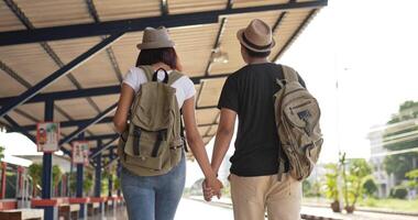 vista traseira do casal feliz viajante asiático com chapéu e mochila mão juntos enquanto caminhava na estação de trem. sorrindo jovem e mulher olhando a vista. conceito de férias, viagens e hobby. video