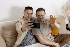 Happy gay couple with casual clothes holding smartphone and talking with friends online, having video call. Homosexual relationships and alternative love. photo