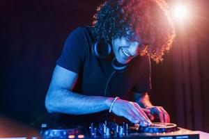 Man with curly hair using DJ equipment and standing in the dark neon lighted room photo