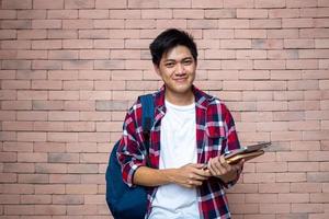 Asian male students wear plaid shirts. Standing next to a brick wall, carrying a backpack, carrying books, school supplies, preparing for study, smiling. photo