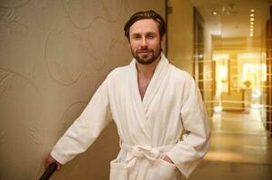 Relaxed handsome middle aged European man dressed in white bathrobe standing on the hallway of a luxury wellness spa resort, smiles toothy smile looking at camera, after getting male beauty procedures photo