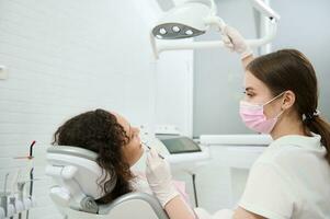 Female dentist in protective medical mask adjusting the lamp and examining the oral cavity of a woman patient sitting in a dental chair in modern dentistry equipped with modern professional equipment photo