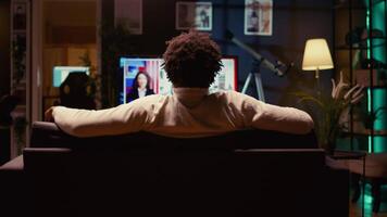 Man holding remote, looking at latest news on TV after zapping through VOD channels on streaming service. Person using modern television set in neon lit living room to entertain himself, zoom out video