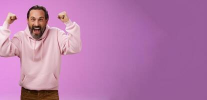 Excited cheering devoted male bearded grey hair yelling supportive words win lottery place good lucky bet standing pleased triumphing say yes raising fists victory success gesture, purple wall photo