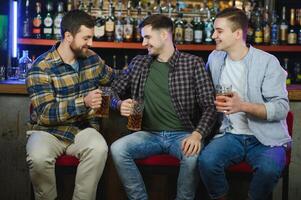 Three young men in casual clothes are smiling, holding bottles of beer while standing near bar counter in pub photo