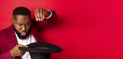 Close-up of charismatic Black male magician perform a trick with his hat, performing on christmas party, standing over red background photo