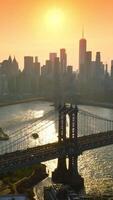 dourado hora pôr do sol Tempo sobre Manhattan arranha-céus. brilhante Sol refletindo dentro leste rio entre dois pontes. laranja céu fundo. vertical video