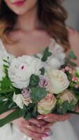 Wedding bouquet of creamy roses in the hands of a beautiful bride. Lovely young woman dressed in white wedding dress is holding flowers. Close-up. Vertical video