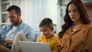 Caucasian family man woman father mother parents working remote online on laptop kid boy child son playing on mobile phone smartphone on couch home gadget addiction modern technology weekend together photo