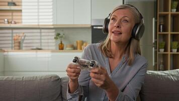 Funny smiling mature caucasian woman playing game console in headphones using joystick controller at home on couch elderly female play computer games older people and modern technology concept photo