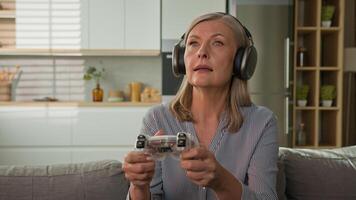 Funny smiling mature caucasian woman playing game console in headphones using joystick controller at home on couch elderly female play computer games older people and modern technology concept photo