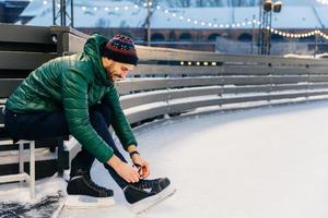 foto de modelo masculino habilidoso com expressão feliz amarra patins, vestido de anoraque verde, senta-se no anel de gelo, vai se envolver em seu hobby favorito. homem se diverte e entretenimento ao ar livre