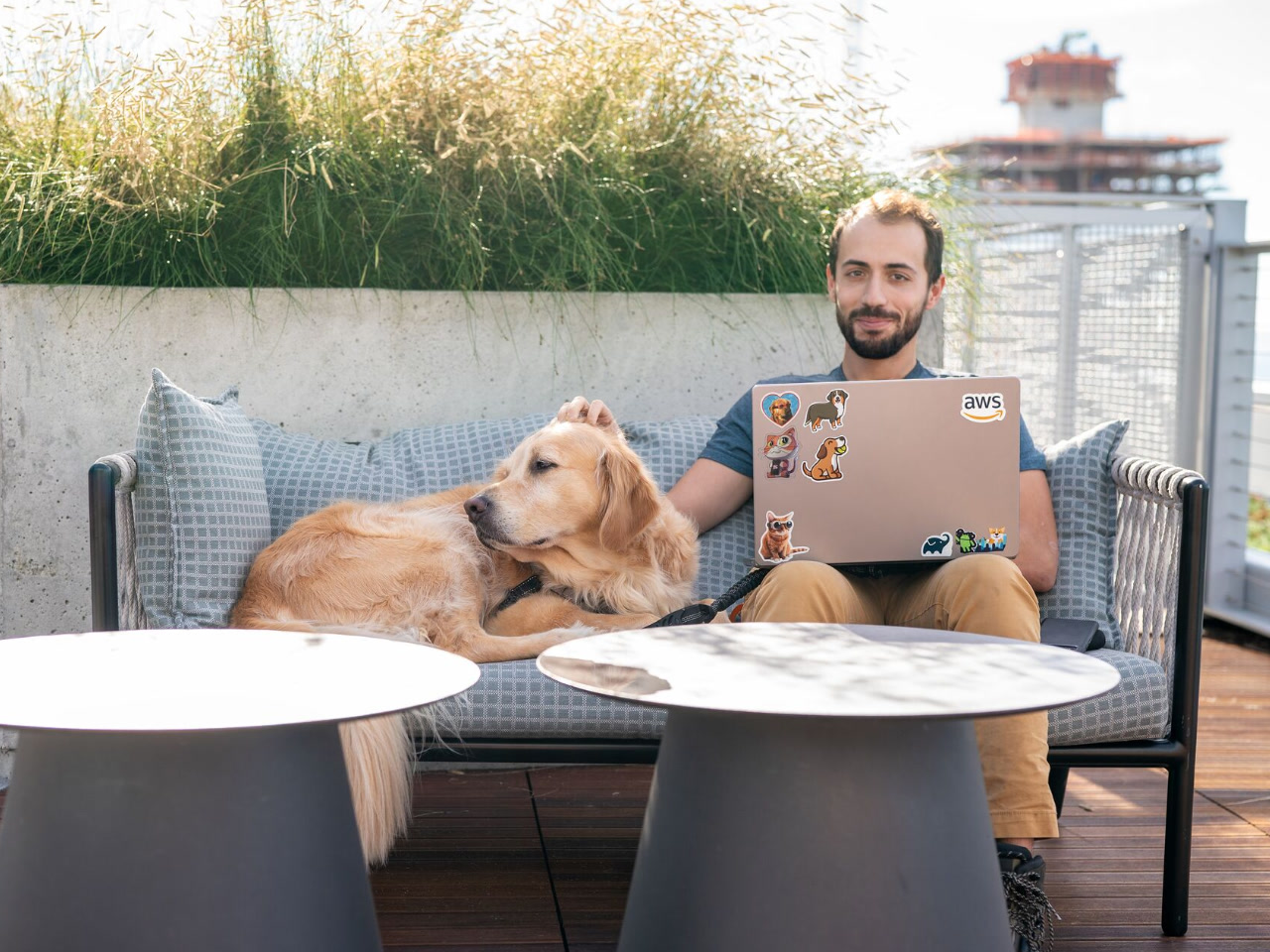 An image of a man seated at an outdoor table at an Amazon office, with his laptop in front of him and a large yellow dog by his side. The man is petting the dog and looking toward the camera.