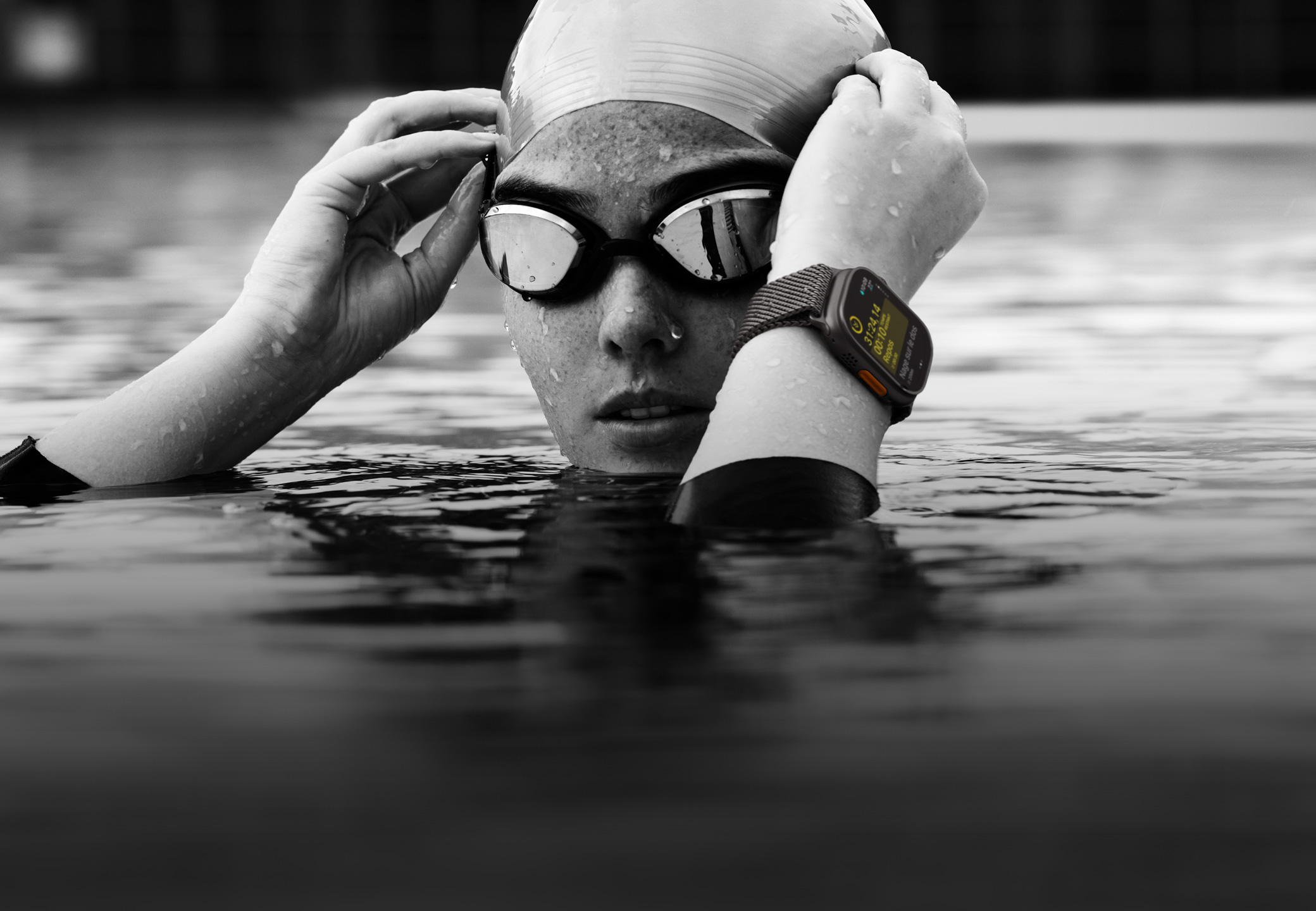 Une personne dans une piscine dont seules la tête et les mains dépassent de l’eau. Elle porte une Apple Watch Ultra 2 et ajuste ses lunettes.