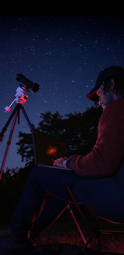 A person outside at night using a MacBook Pro on their lap, connected to a device on a tripod pointed toward the starry sky.