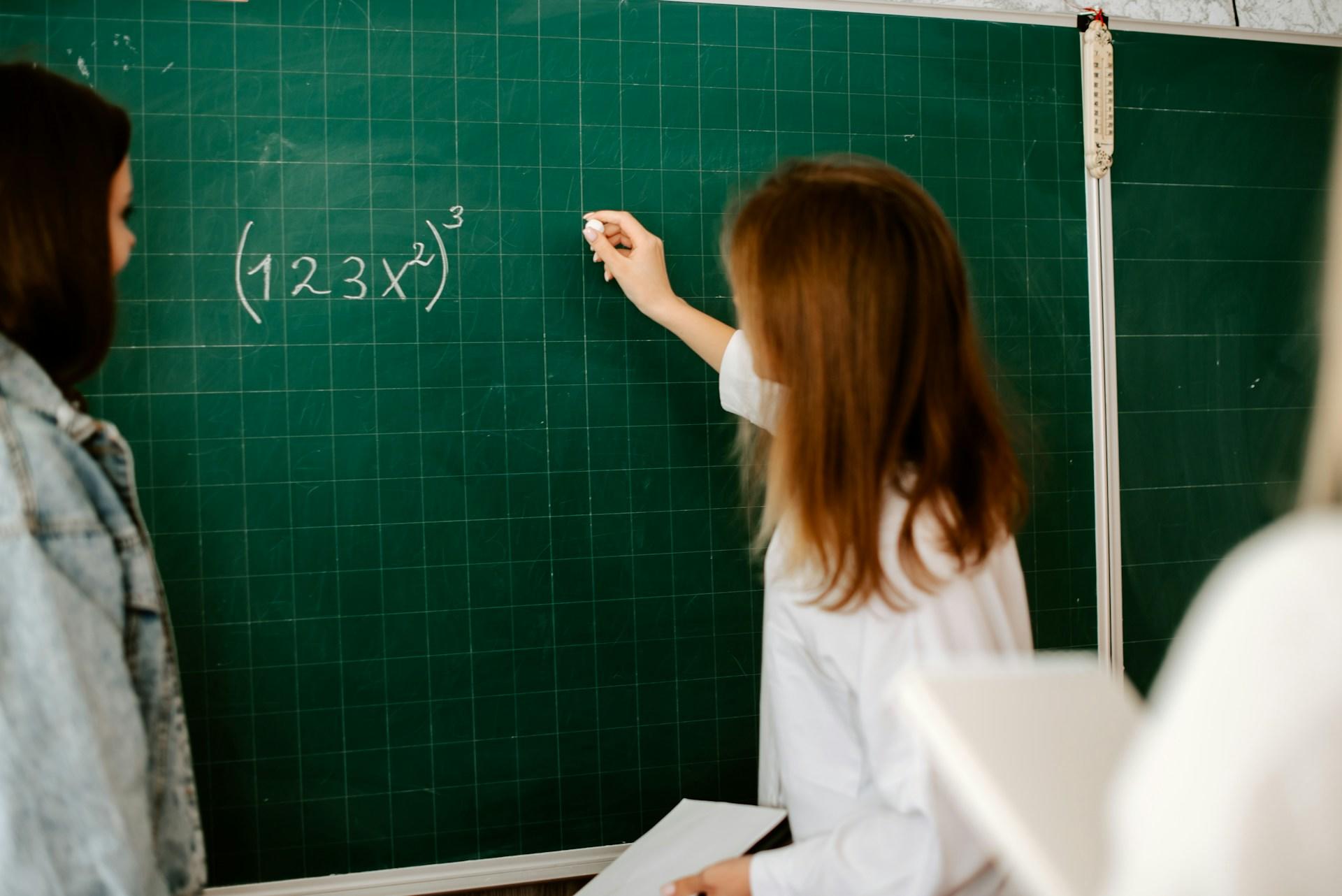 Eine Person schreibt eine Mathe Aufgabe an die Wandtafel und zwei Personen schauen dabei zu.