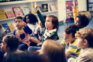 Photo of a group of children sitting on the floor in a classroom, looking n one direction, several with their hands up.