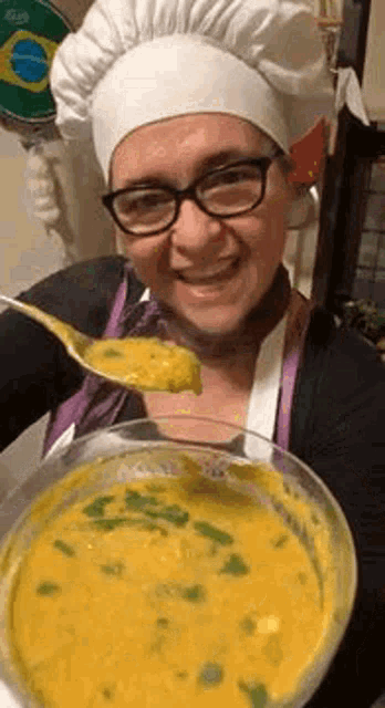 a woman wearing a chef 's hat and apron is holding a bowl of soup and a spoon in her mouth .