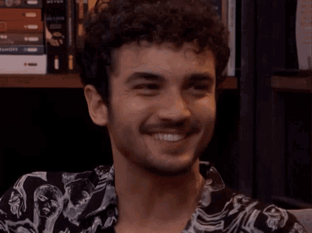 a man with curly hair and a beard is smiling in front of a bookshelf with books on it .