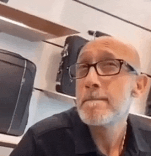 a bald man with glasses and a beard is sitting in front of a shelf with suitcases .