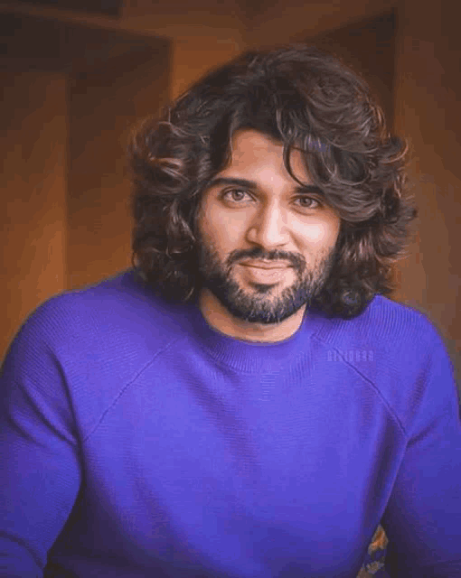 a man with long curly hair and a beard is wearing a blue sweater and smiling for the camera .