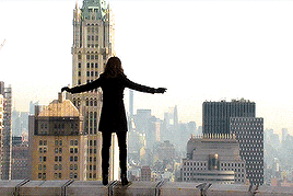 a woman is standing on the edge of a building with her arms outstretched in front of a city skyline .