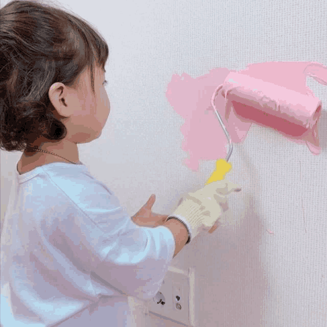 a little girl is painting a wall with a pink paint roller