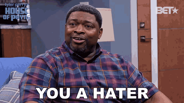 a man in a plaid shirt is sitting on a blue couch and says " you a hater "