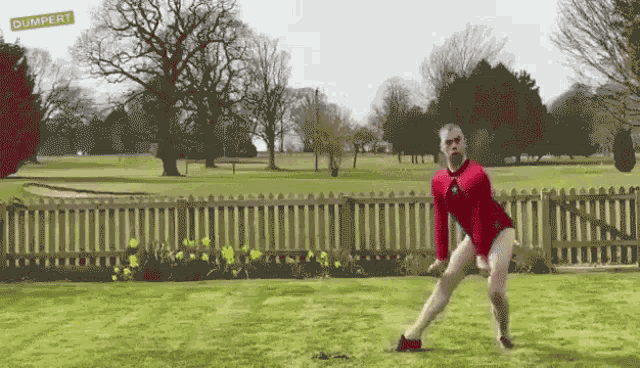 a man in a red shirt and shorts is standing in a grassy field with a dumpert sign in the background