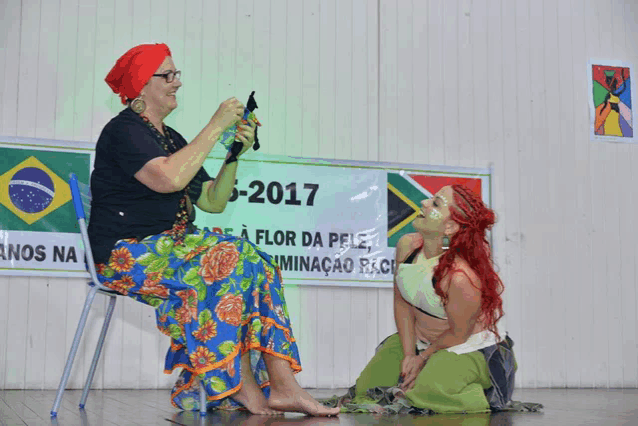 a woman sits in a chair next to a woman kneeling in front of a sign that says " 2017 "