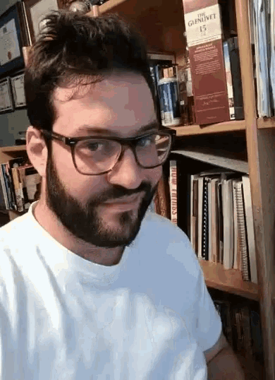 a man with glasses and a beard is standing in front of a bookshelf with a bottle of gunnive whisky