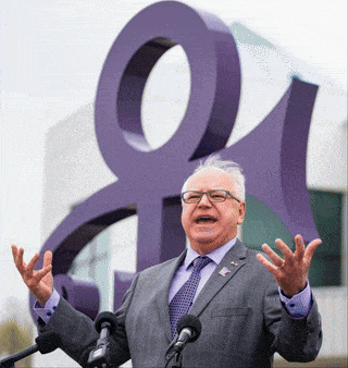 a man in a suit and tie stands in front of two microphones in front of a large purple symbol