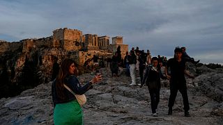 Touristen spazieren auf dem Areopag-Hügel vor der antiken Akropolis, während die Sonne in Athen untergeht.