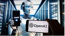 لوگوی OpenAI روی صفحه یک تلفن همراه در مقابل تصویری بر روی صفحه یک کامپیوتر که توسط مدل تبدیل متن به تصویر Dall-E  ایجاد شده است،