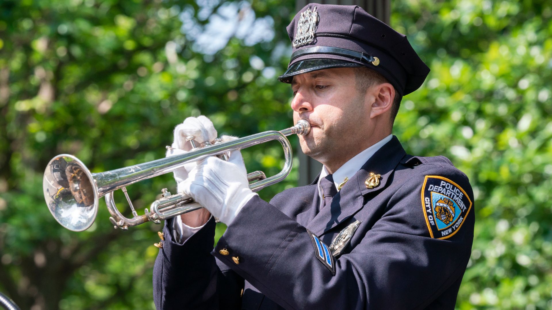 A man in a navy blue uniform is seen playing a silver horn, with white gloves on, against a backdrop of green tree leaves