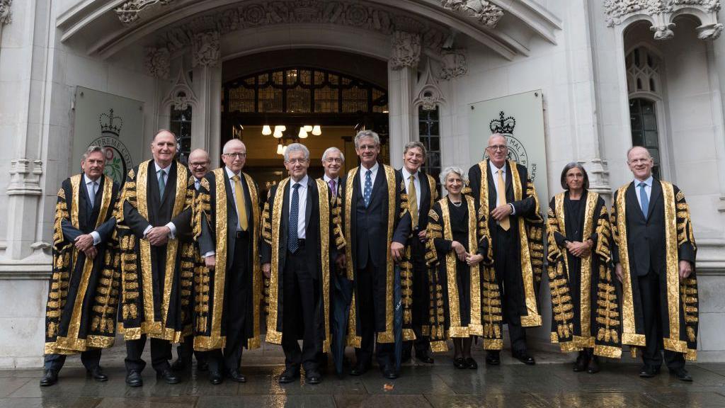 The judges of the Supreme Court gathered outside the doors. There are 10 men and two women standing in a row wearing the black and gold robes of the court.