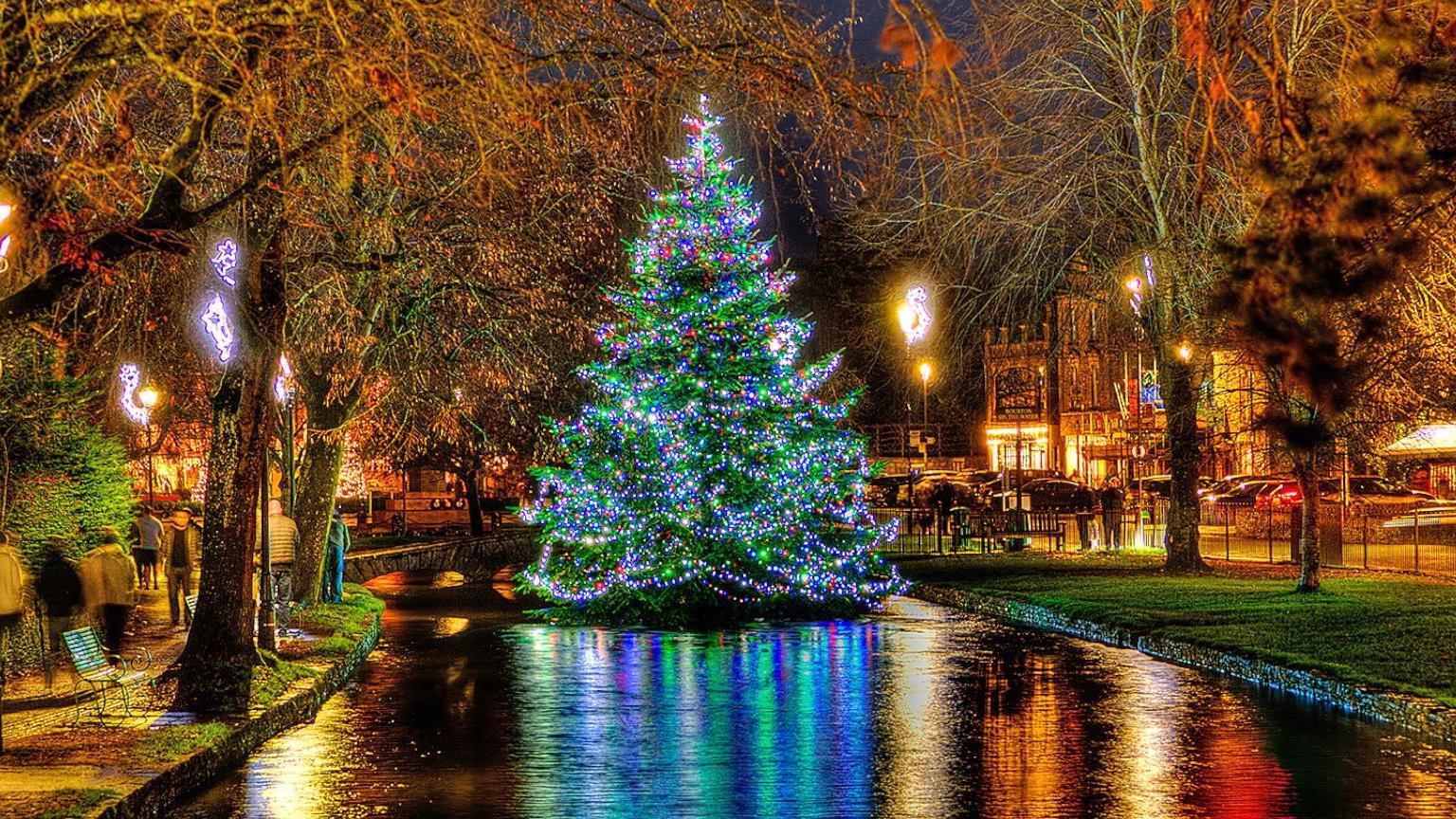 A Christmas tree in the river in Bourton-on-the-Water lit up at night. The tree's colourful blue, green and pink lights are reflected in the river, while yellow, orange and red lights from near by shops are also picked up in the water. People are walking along the river bank and there is a small low bridge behind the Christmas tree. 