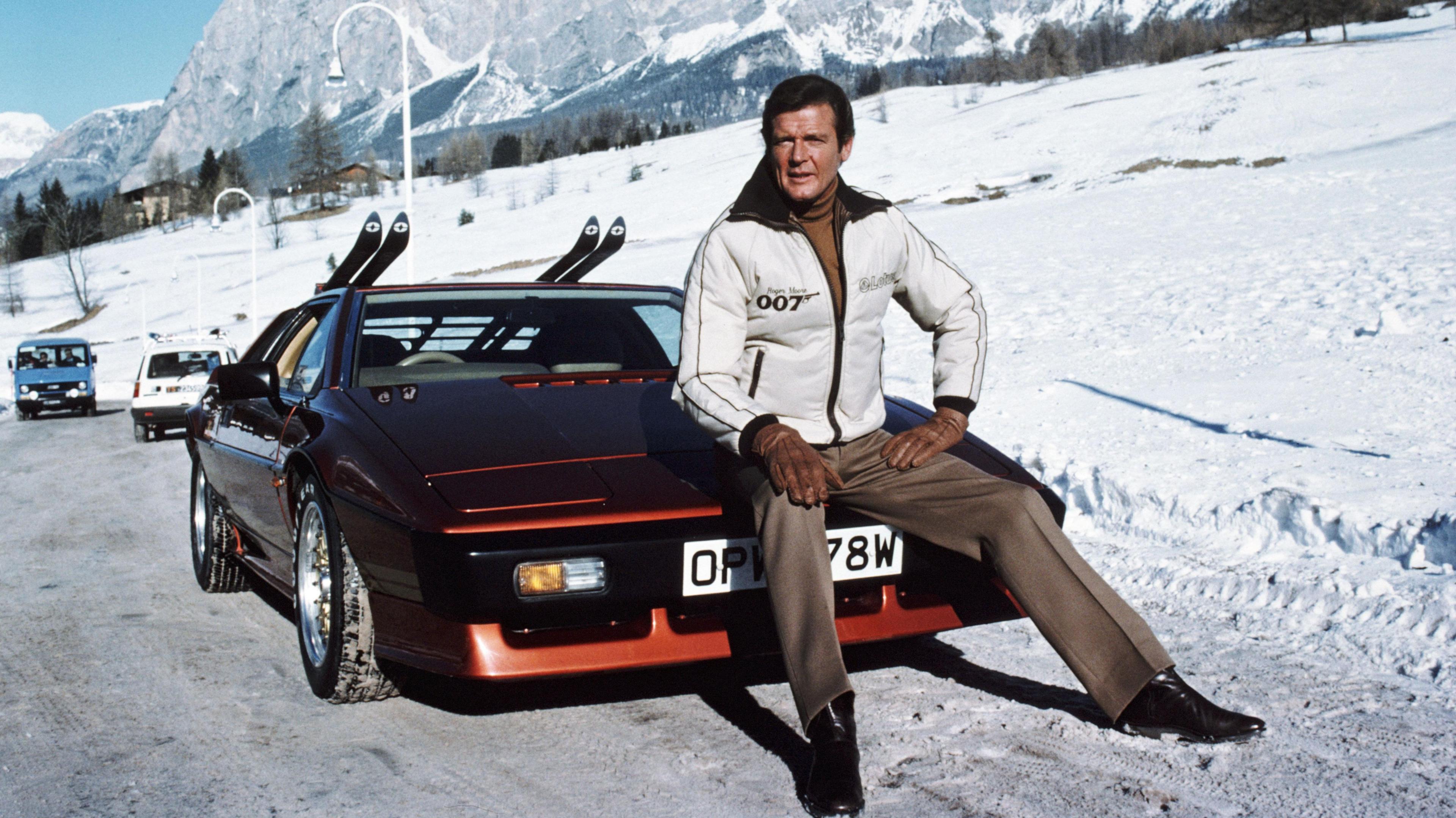 Sir Roger is sitting on the bonnet of a black and red Lotus Esprit Turbo sports car. Behind him are snow covered mountains. Sir Roger is wearing a warm white puffy jacket, brown trousers and brown gloves. The numbers "007" can be seen written on his jacket. On the back of the car, are two sets of skis.