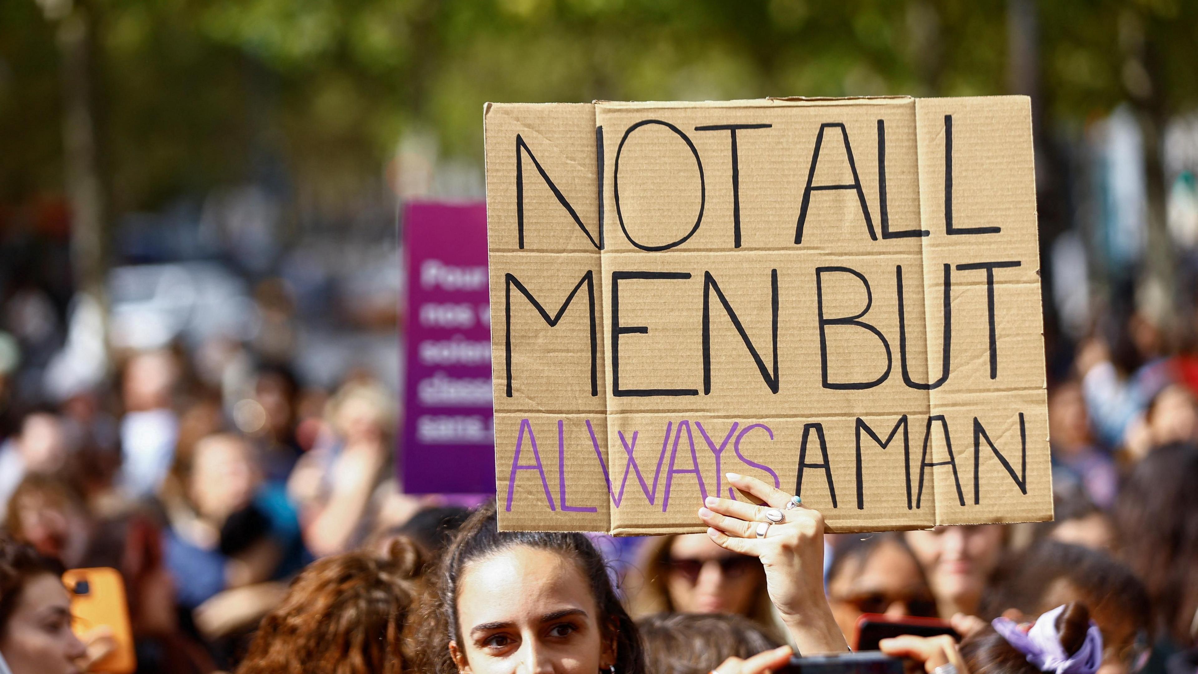 A woman holds a sign saying Not All Men but Always a Man