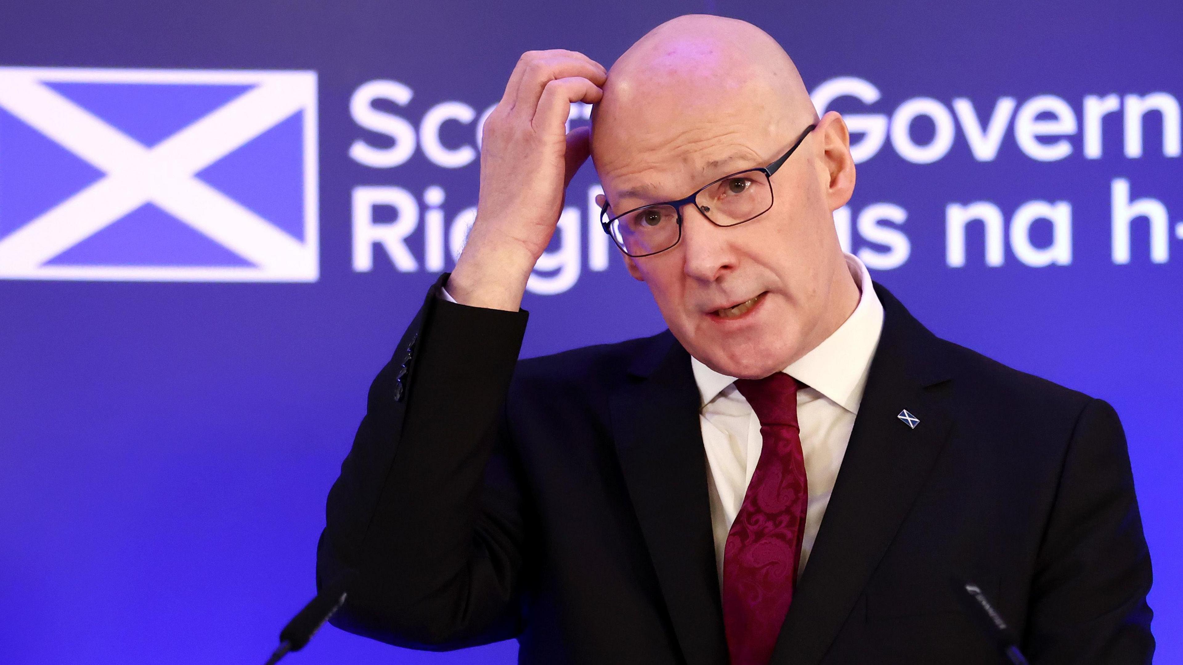 John Swinney, first minister of Scotland, scratching his head at a press conference. He is weating a dark suit, white shirt and burgundy swirly tie. He is wearing an enamel saltire badgs on his left lapel.