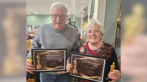 A man and a woman, who are both retired and work as volunteers, stand in a hospital atrium holding framed "Voluntary Oscars" certificates and miniature gold-coloured, Oscar-style trophies. Graham stands on the left in a grey jumper and jeans, while Jean wears a sparkly jumper.