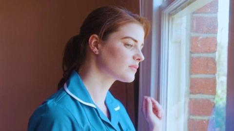 A young midwife with  hair in a ponytail and blue uniform on stares out of a window, looking down, with her hand against the glass in a loose fist.