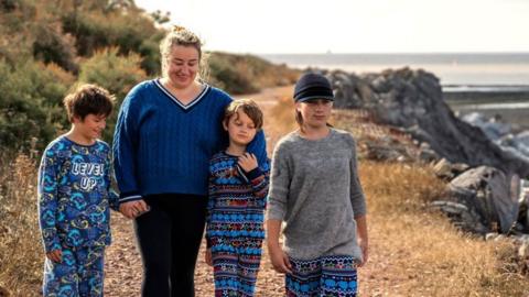 Sammiie wearing blue Converse shoes, black skinny jeans and a blue knitted jumper. She is walking alongside her three children on a coastal path next to the beach, which is full of rocks and shingles. It is a bright but slightly overcast day. All three of the boys are wearing blue patterned pyjamas.