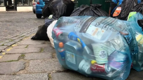 Bin bags mainly black but one see through blue lying on the pathway by a road.