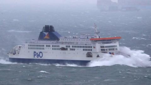 A blue and white P&O ferry in the sea. A large wave is crashing against the back. There are large waves in the water.