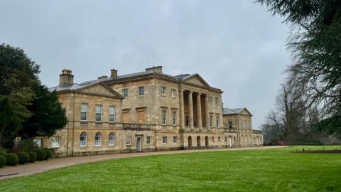 A large country house sits in the centre of the picture. The house has at least three floors with several pillars forming part of its front. It sits in parkland with several trees. The sky is overcast.