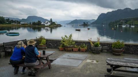 Two people sit a picnic table in Plockton in Wester Ross. They are enjoying coffee and looking out over the village's bay. 