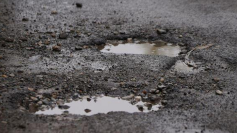 Close up view of two potholes full of water on a black-tarmac road with several stones surrounding them.