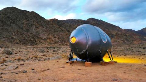 A circular pod is seen parked in the mountains of Ladakh, it is sliver in colour and held up with little pole structures held in place with plywood boxes, with the browny-red backdrop of the Ladakh mountains in the background and some clouds in a dark sky.